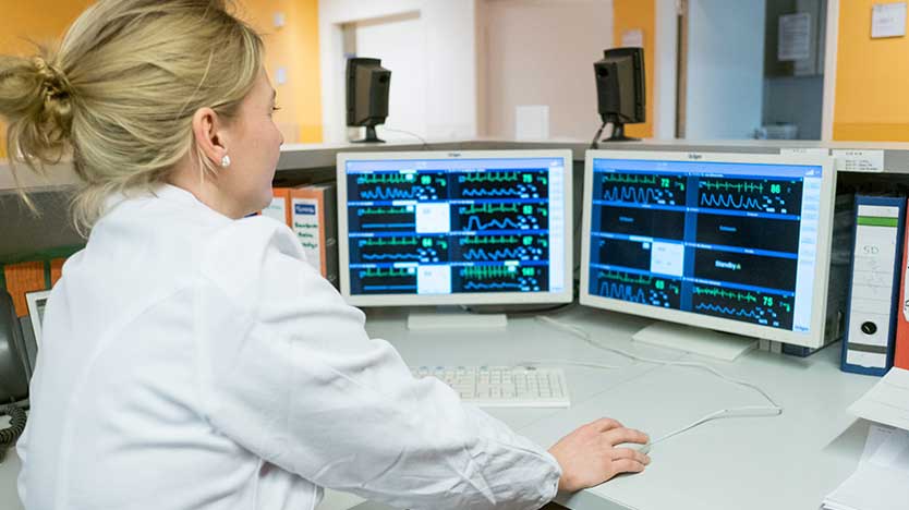 Ärztin betrachtet zwei Monitore mit bunten Kurven und Zahlen auf schwarzem Grund; Quelle: BMWi / Ole Spata