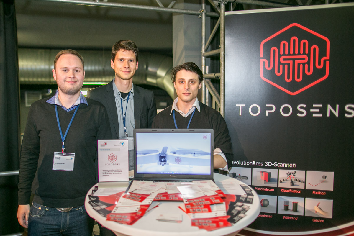 Dieses Bild zeigt die drei Gründer des Start-ups Toposens an einem Informationsstand zu ihrem Unternehmen. 